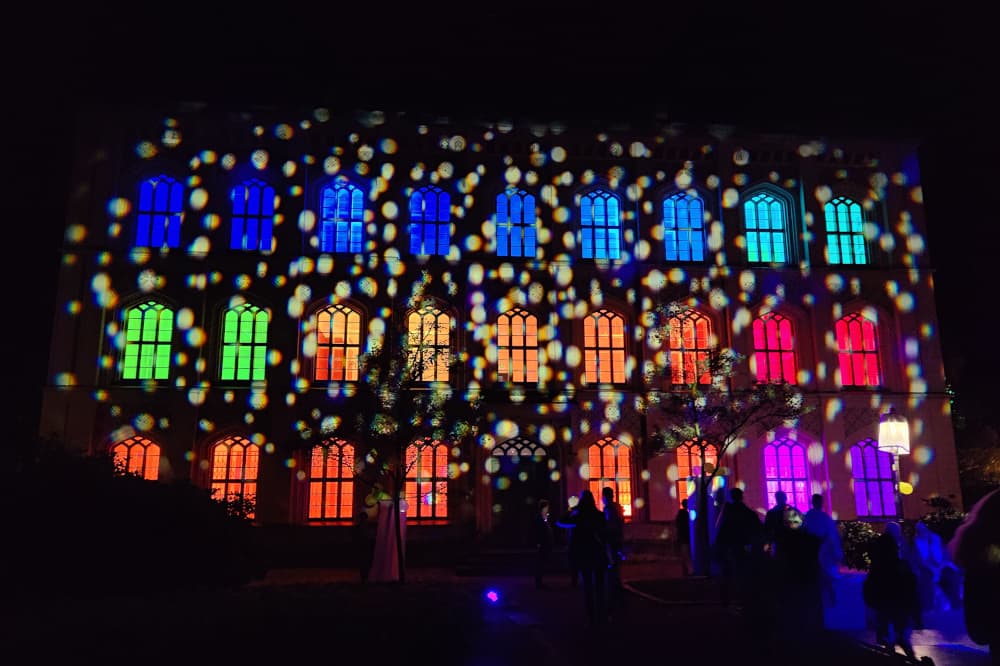 Photo of the Building Trades School at night. The building is illuminated inside with different colored lights and illuminated outside with white dots.